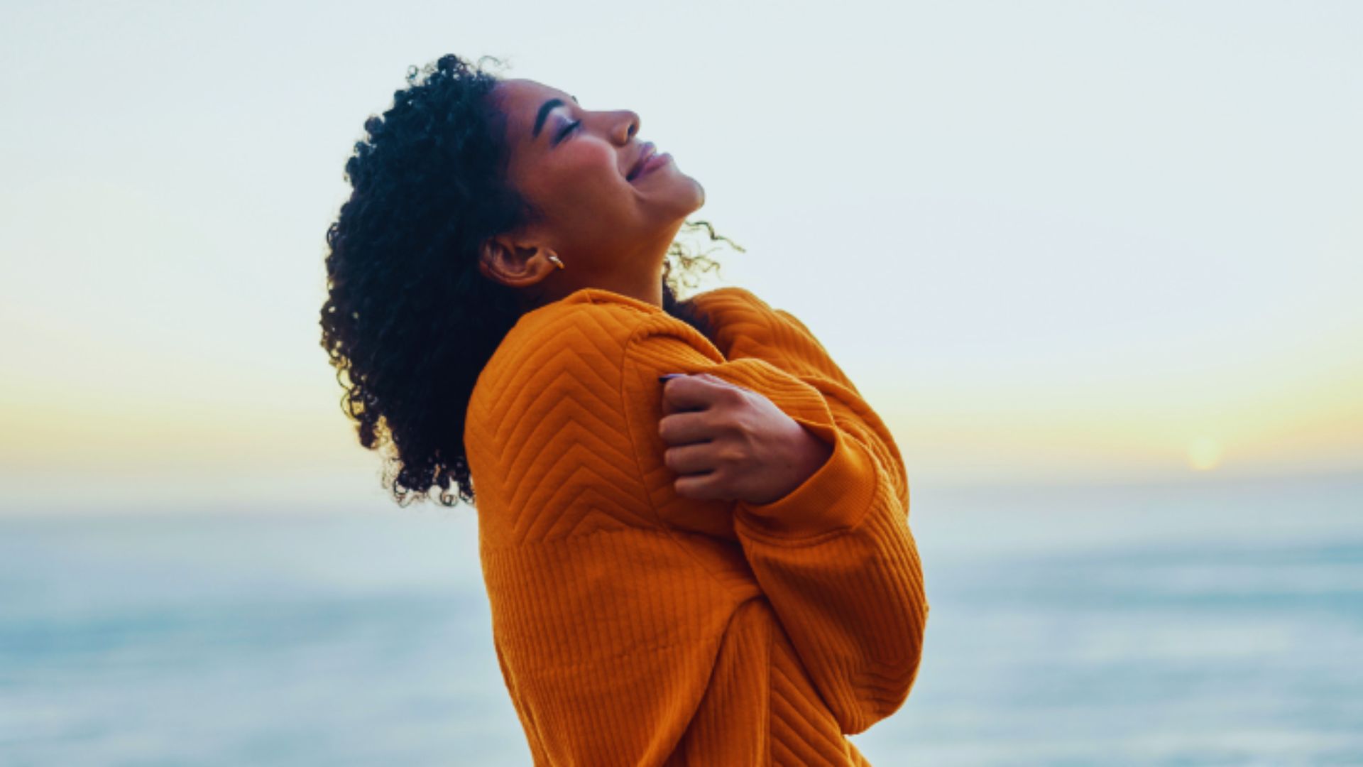 A woman in an orange sweater stands on the beach, gazing at the ocean waves under a clear blue sky.