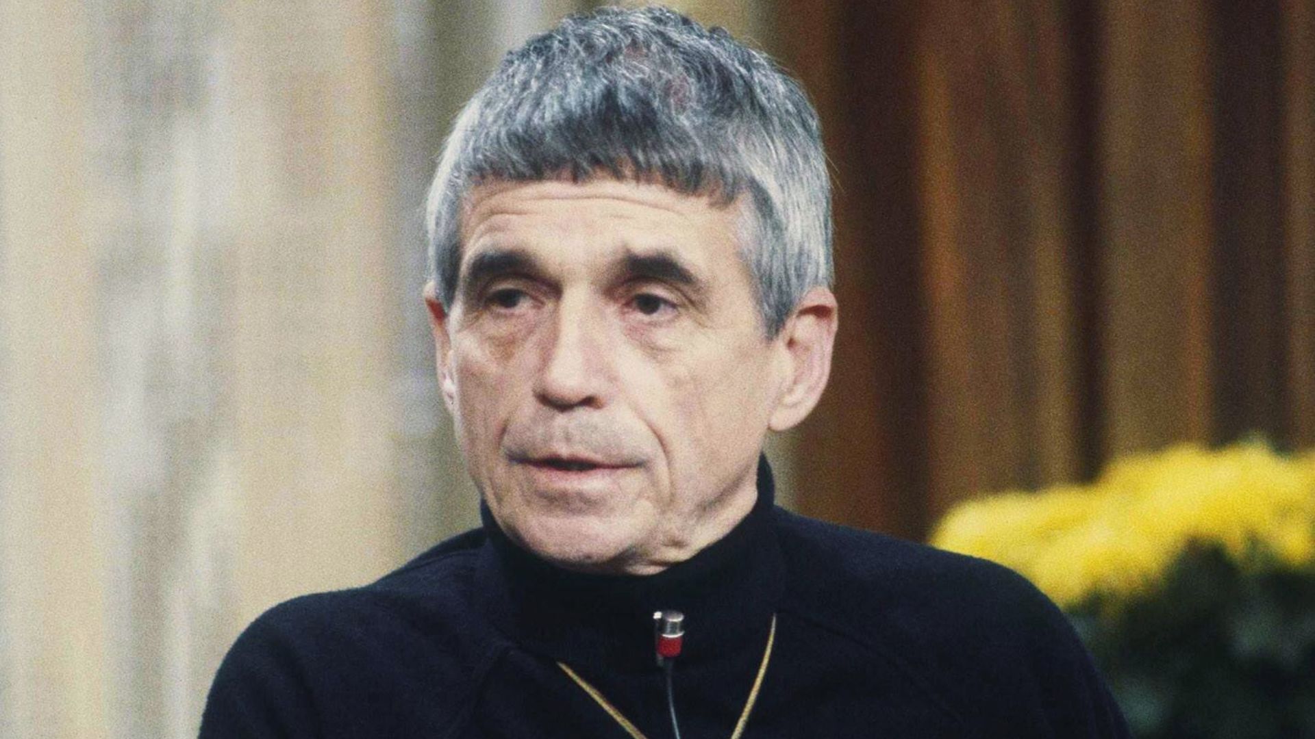 A man with grey hair wearing a black shirt, looking directly at the camera with a neutral expression.