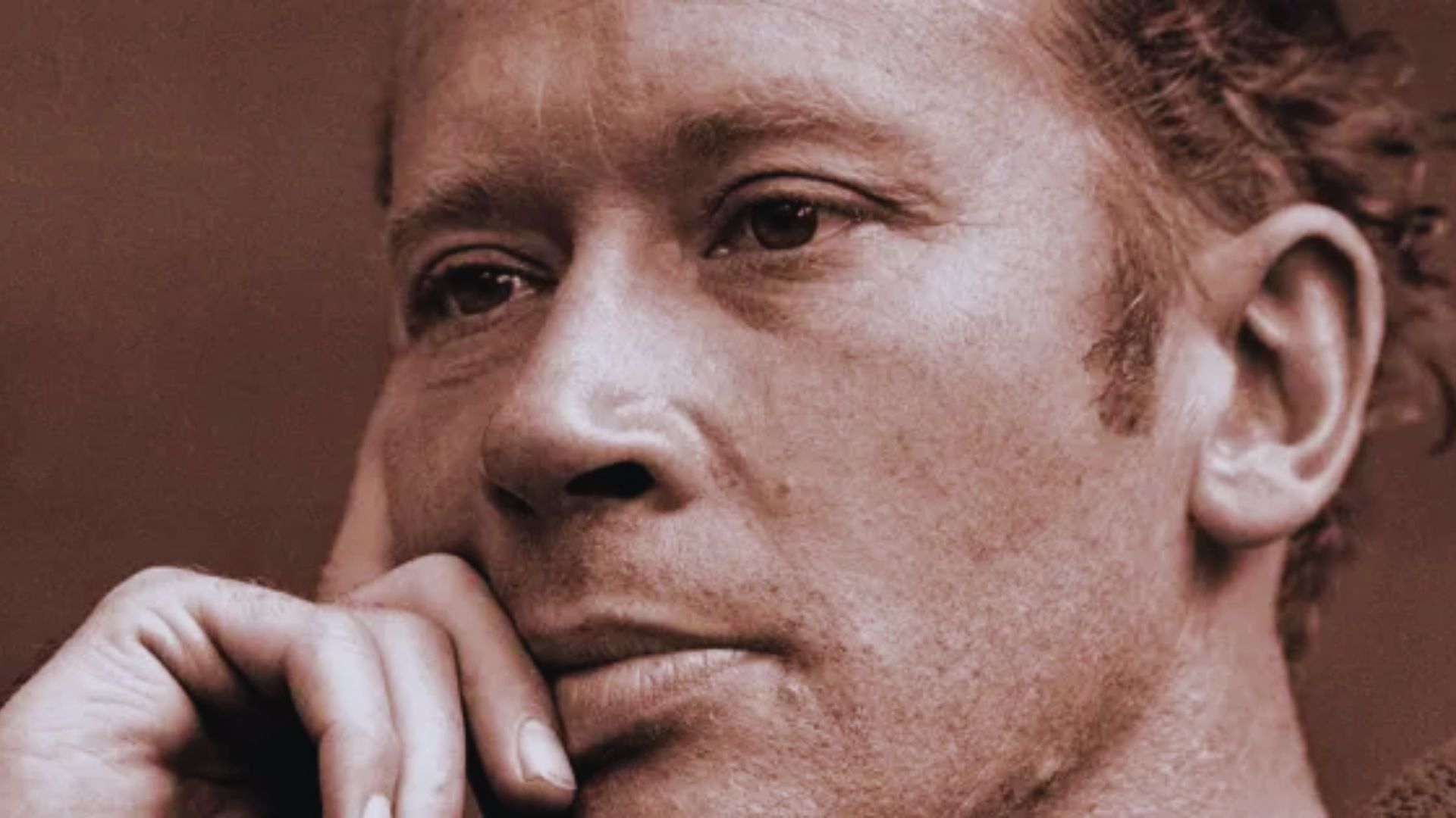 A man with a pensive, sad expression, his hand resting on his chin.