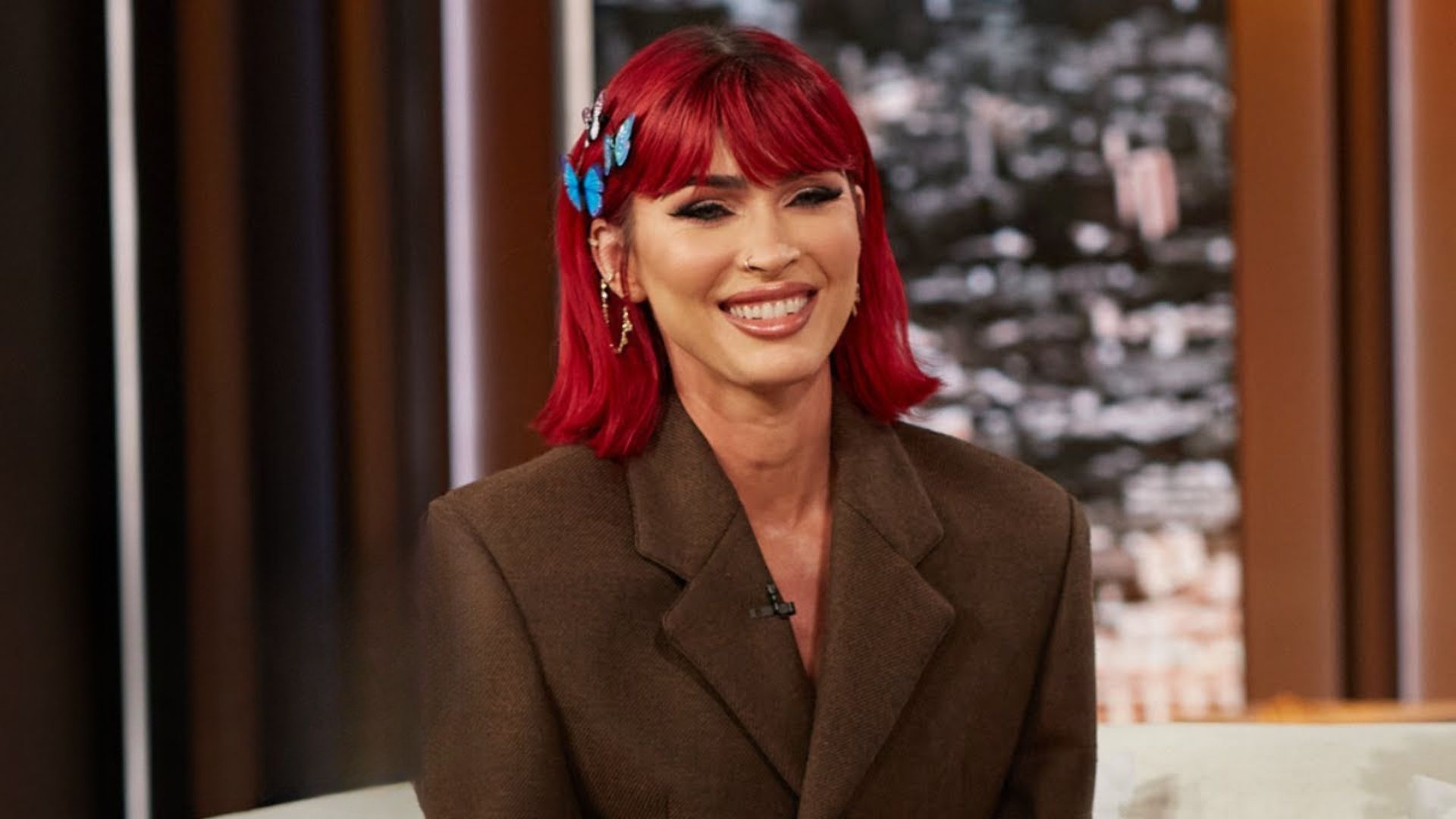 A woman with red hair wearing a brown jacket stands confidently, showcasing her vibrant hair against the neutral jacket.