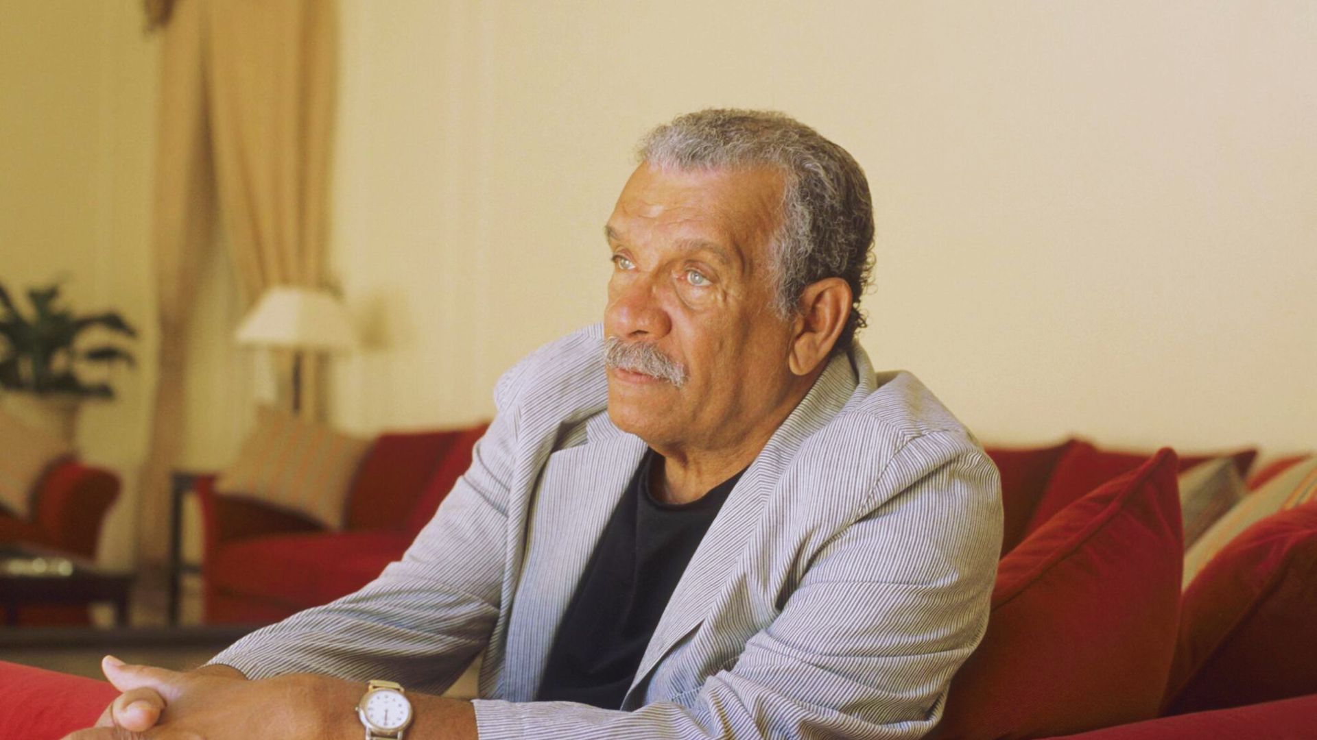 An older man sitting comfortably on a red couch, looking relaxed and engaged in thought.