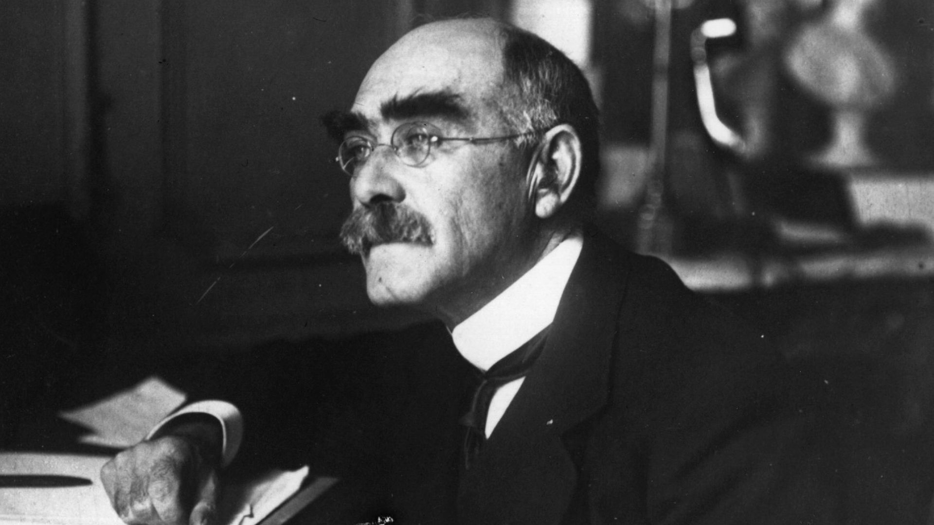 A man with glasses and a mustache is seated at a desk, focused on his work.