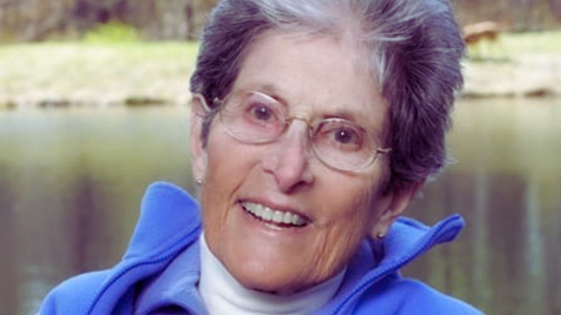 An older woman with glasses and a blue jacket smiles warmly at the camera.