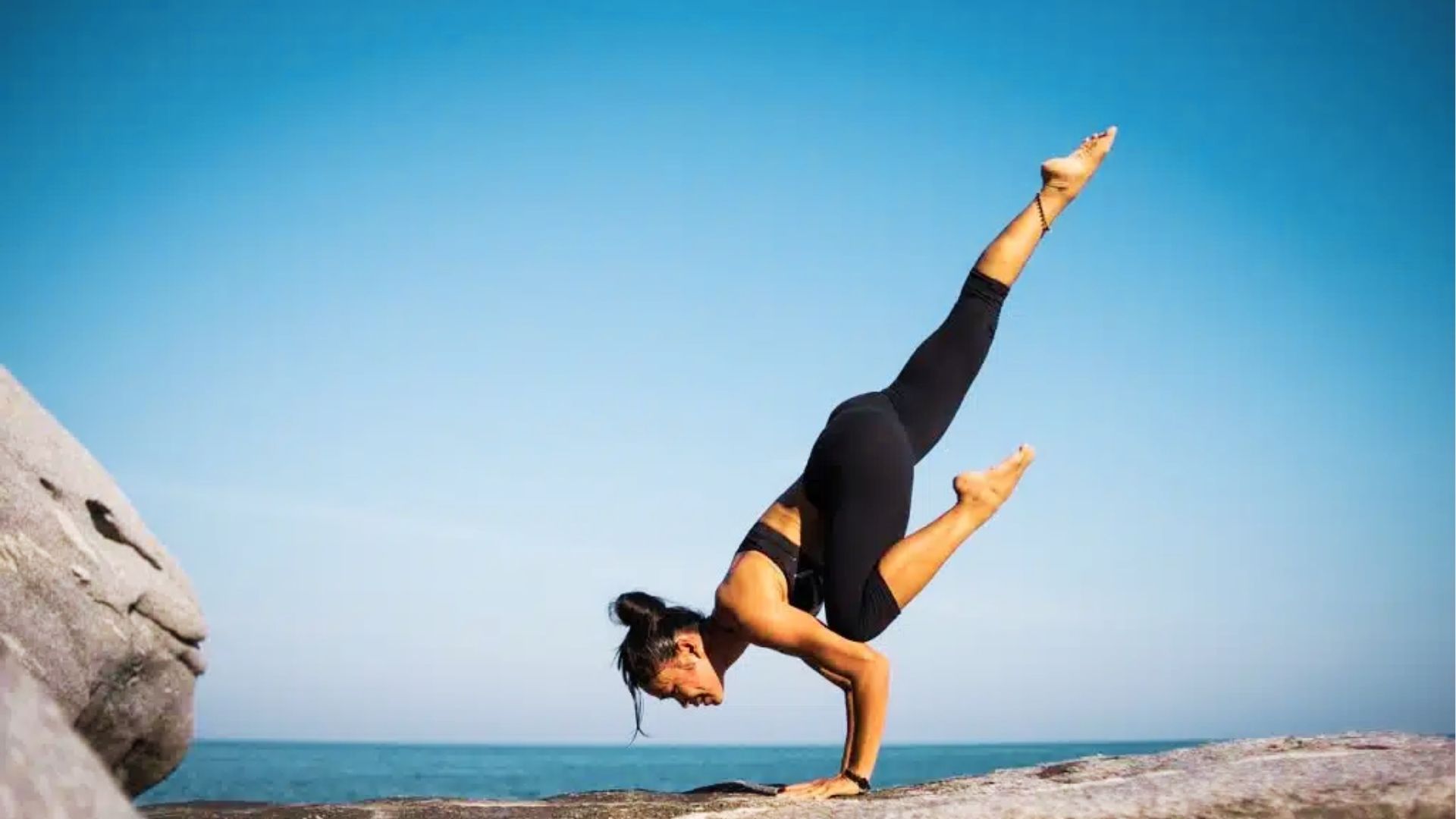 A woman holds a powerful arm balance on sunlit rocks, one leg lifted high against a vast blue sky and ocean.