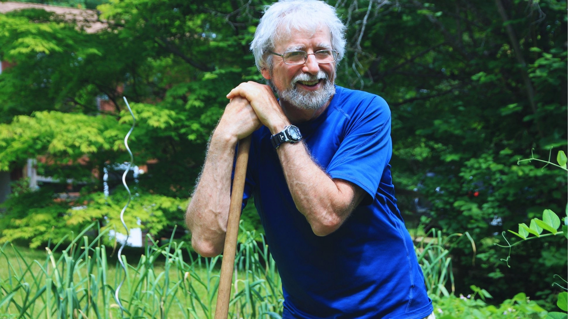 Smiling Tom Wayman poet with white hair and beard in blue shirt leaning on garden tool in lush green backyard.