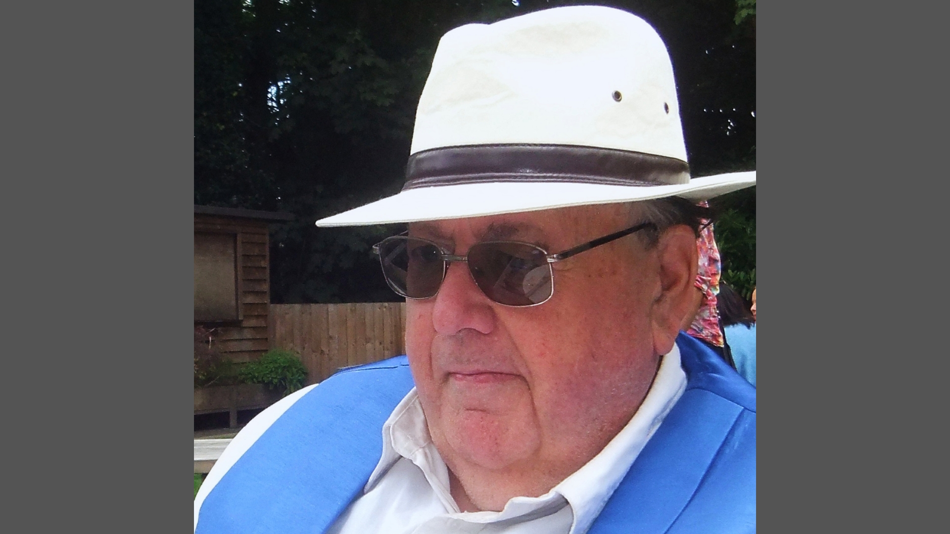 Poet Colin Ian Jeffery wearing white hat, sunglasses, and blue vest outdoors with wooden fence and green trees behind him.