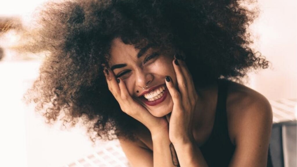 A woman with curly hair cradles her face in her hands, eyes closed in a radiant, unguarded laugh.