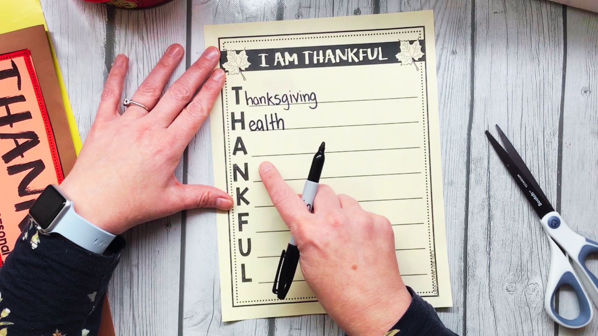 Hands filling out an I AM THANKFUL acrostic poem worksheet on a white wood desk with scissors and craft supplies nearby.