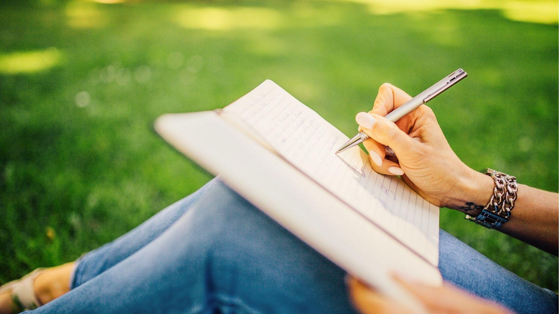 Person writing poetry in notebook outdoors on grass, capturing natural inspiration and the calming rhythm of creative expression.