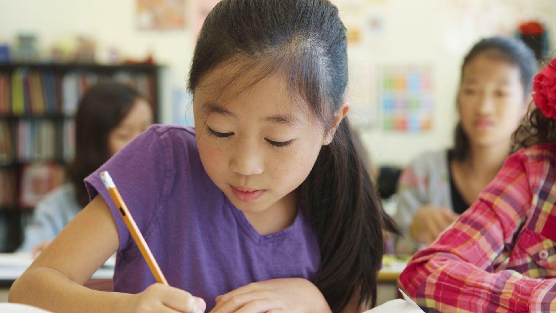Young girl in purple shirt writes with pencil in classroom while classmates sit behind her during creative writing lesson.