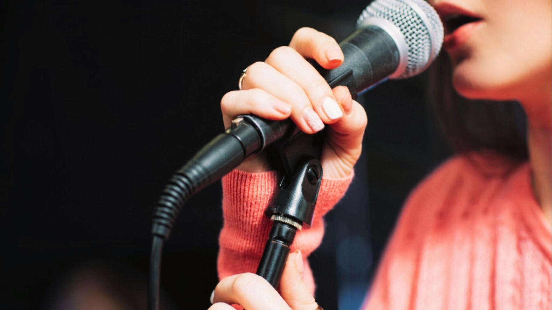 Poet in coral sweater performing spoken word into microphone on stage, practicing delivery and voice projection.