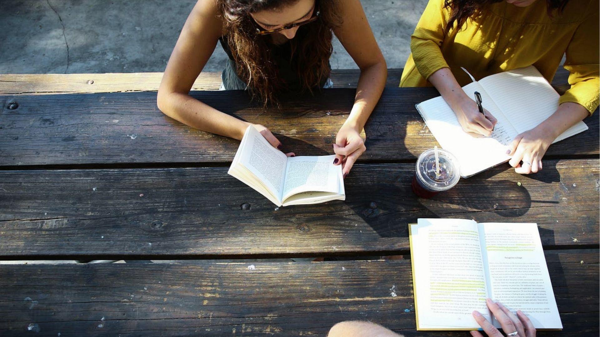 Two writers at a wooden table with notebooks and books, brainstorming and editing spoken word poetry pieces together.