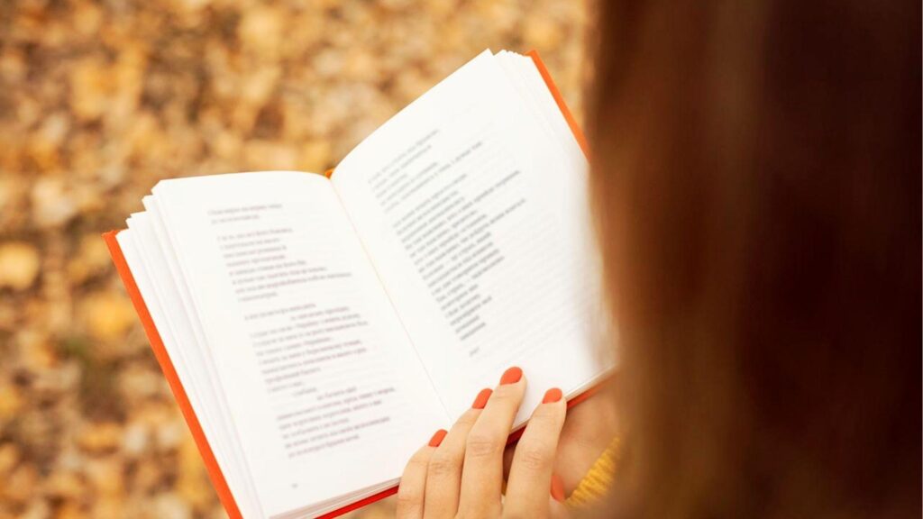 Student writing poetry analysis essay in notebook with open textbook on wooden desk for literature study guide