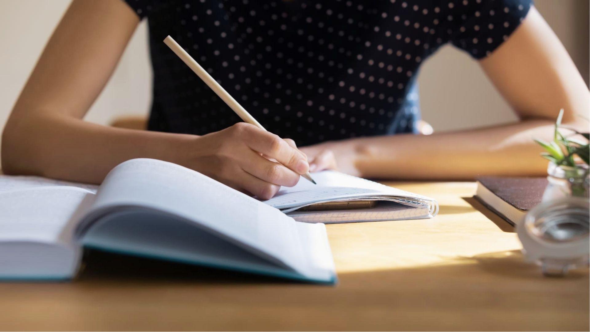 Student writing poetry analysis essay in notebook with open textbook on wooden desk for literature study guide