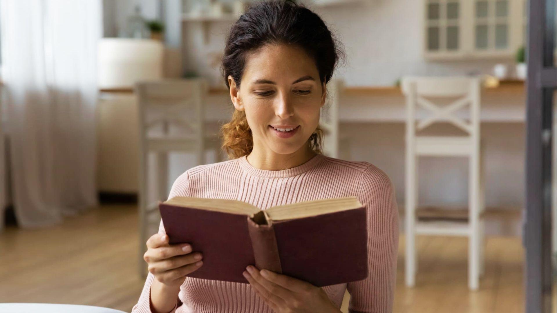 Smiling woman in pink sweater reading poetry book at home, representing literary analysis and essay writing inspiration