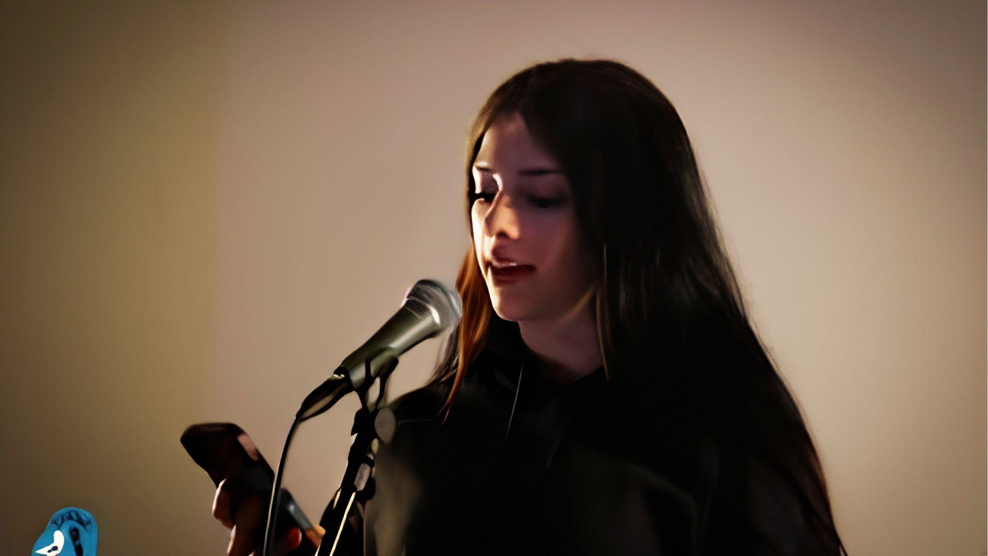Female spoken word poet holding phone and performing into mic, using notes to deliver emotional poem live.