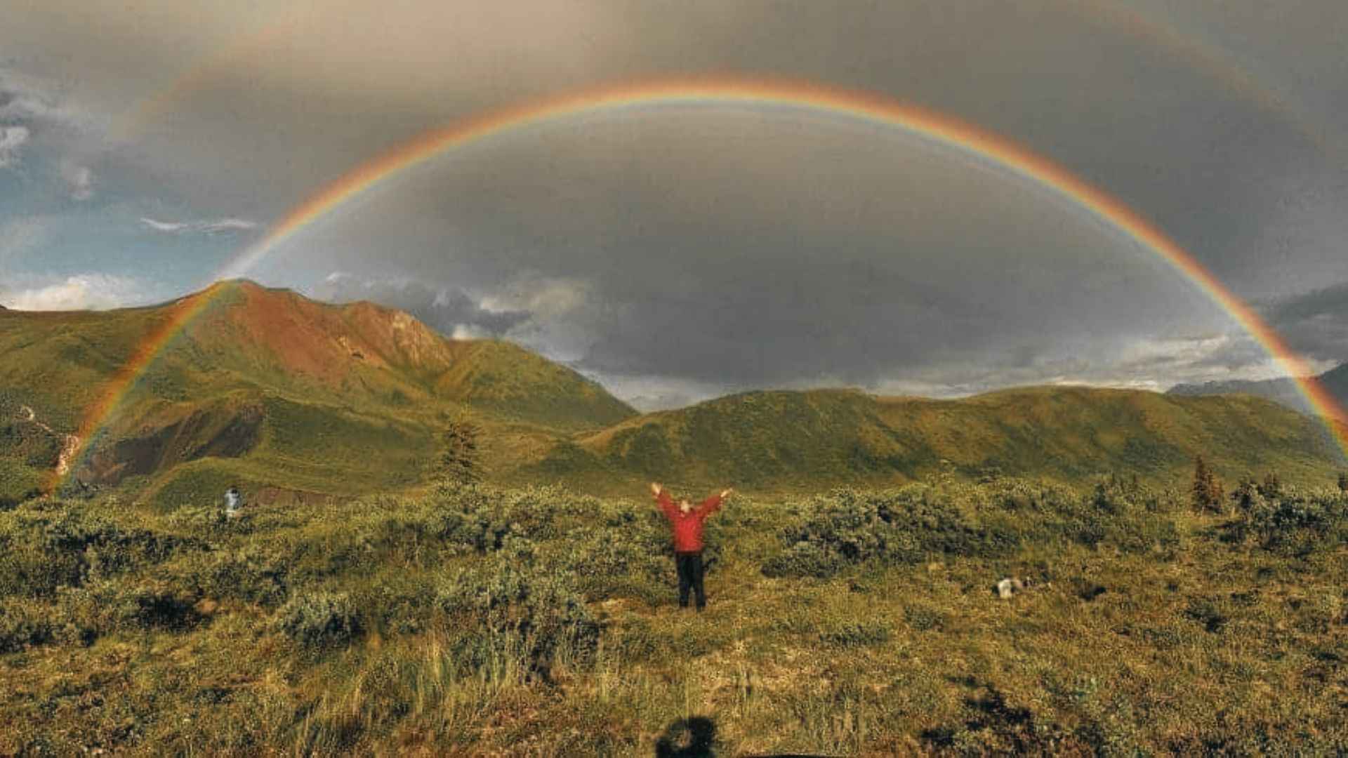 A person stands with arms raised under a vivid double rainbow across mountain slopes.