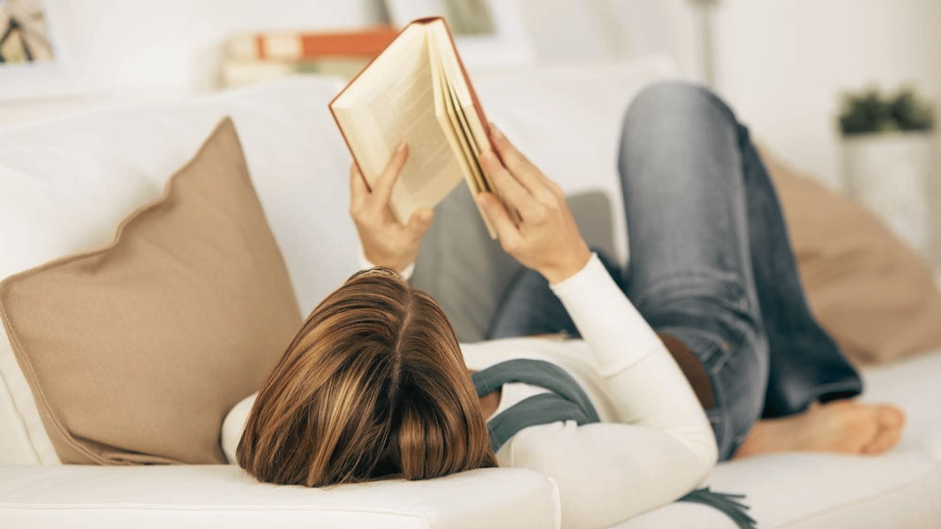 A woman reclines peacefully on a couch, lost in a book she holds above her.