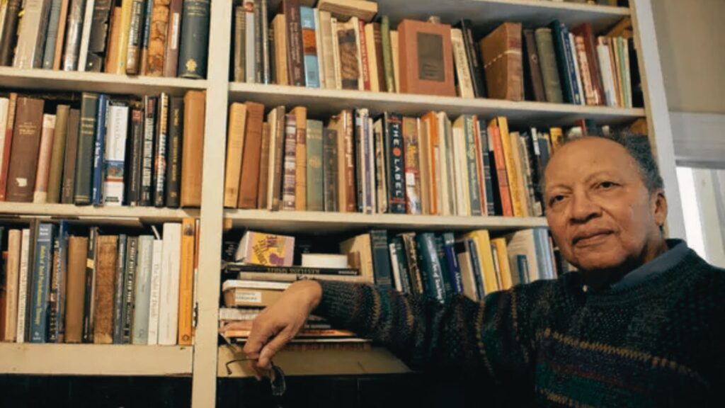 Walter Dean Myers sits beside floor-to-ceiling bookshelves filled with books, holding glasses and wearing a patterned sweater.