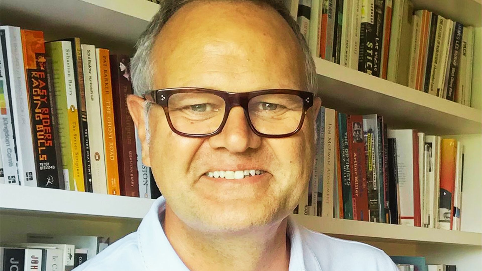A smiling man with gray hair and glasses stands before bookshelves filled with novels.