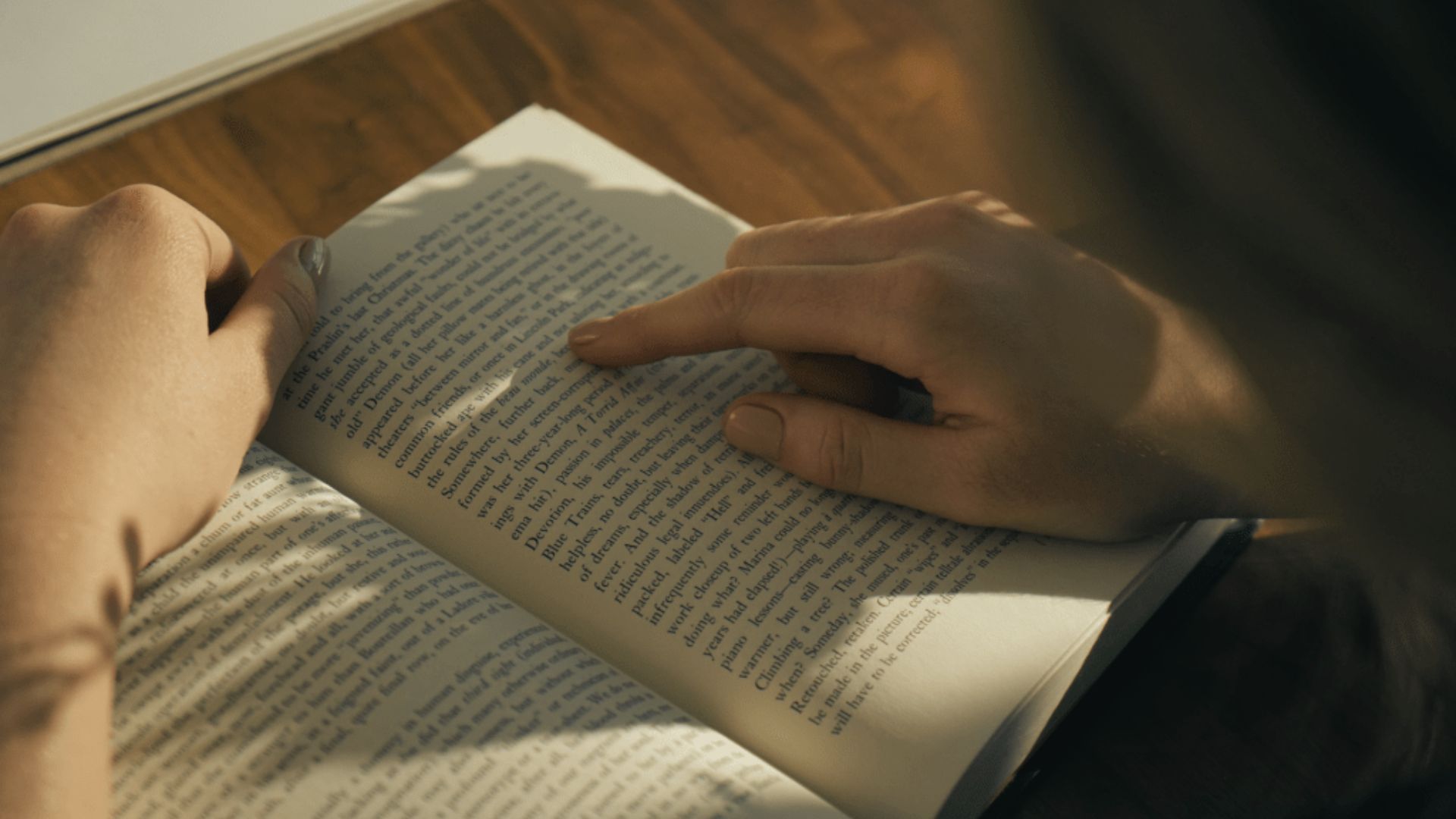Close-up of hands turning pages of an open book in warm sunlight, with one finger pointing to text.