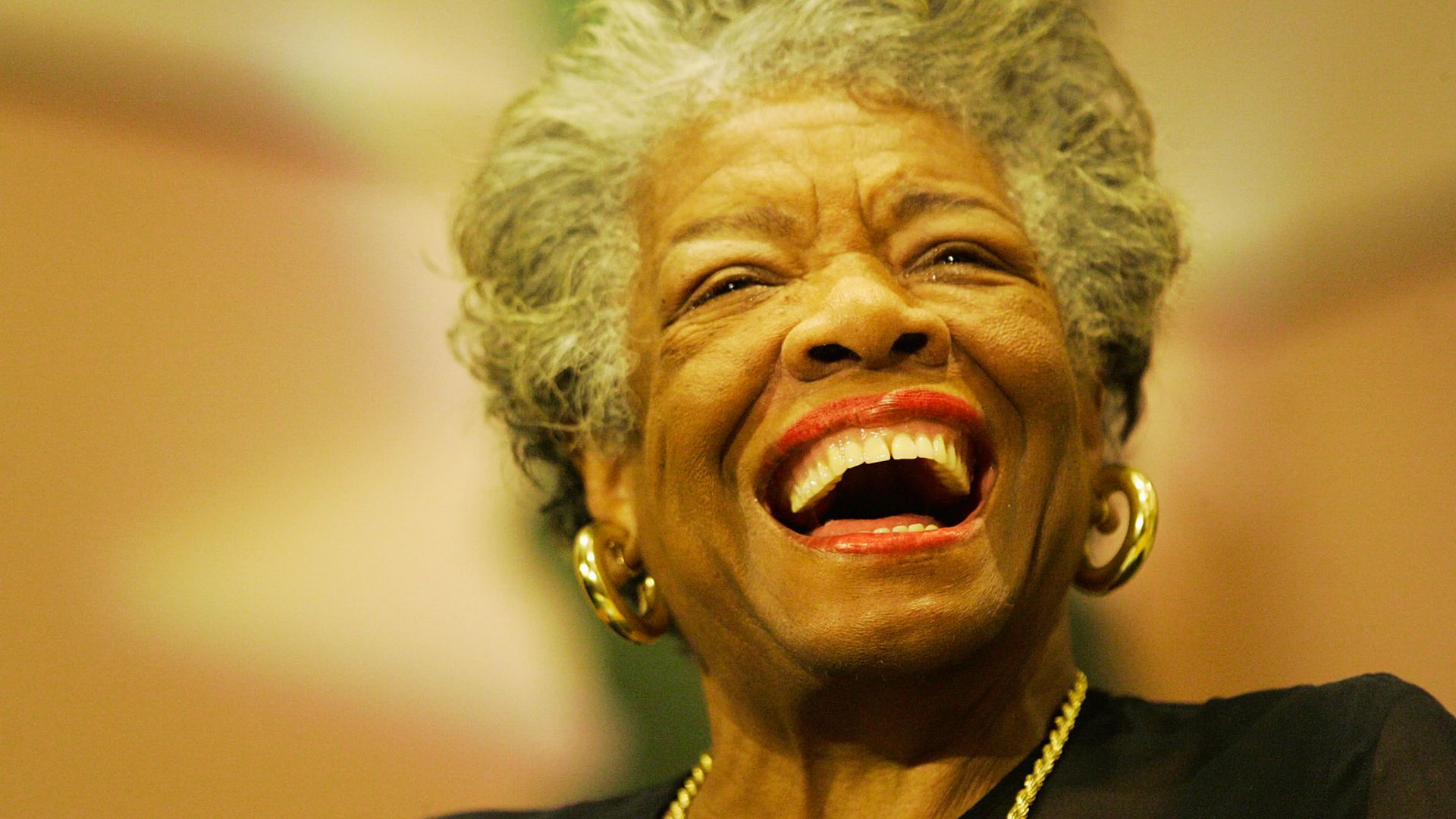 Close-up of Maya Angelou laughing joyfully with red lipstick and gold hoop earrings.