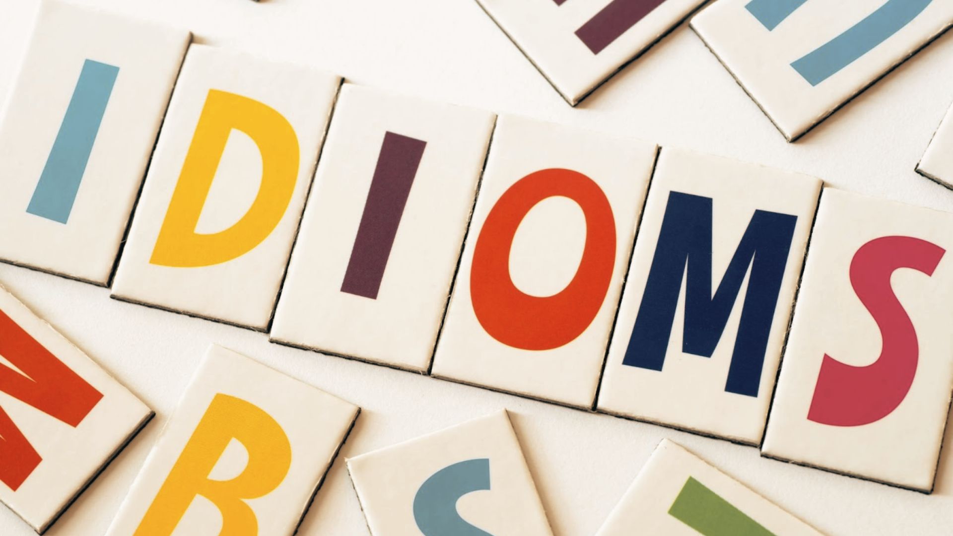 Colorful letter tiles spell “IDIOMS” on a white background with scattered letters around it.
