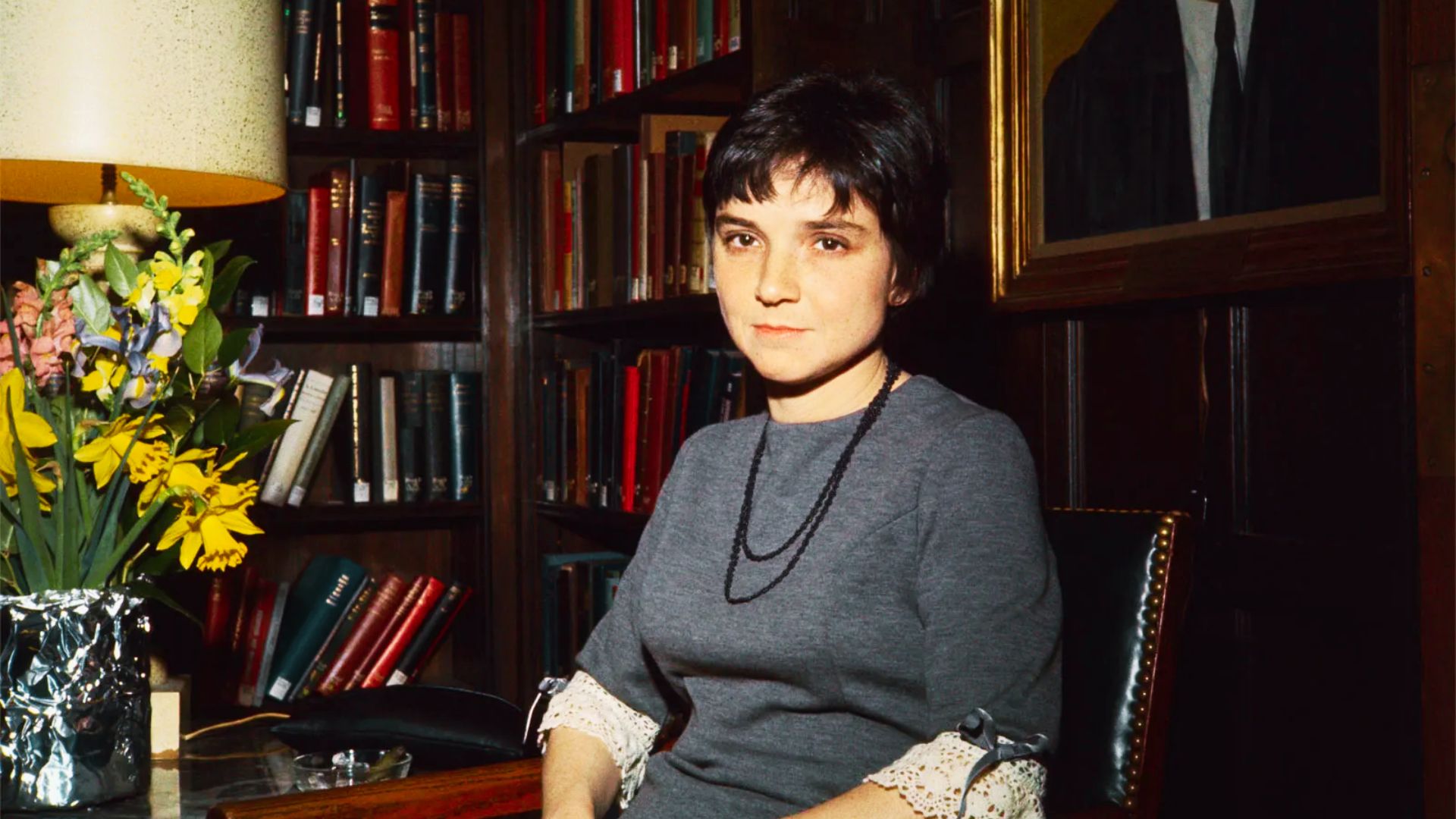 Poet Adrienne Rich sits in a wood-paneled library wearing a gray dress with lace cuffs, beside yellow daffodils and bookshelves.