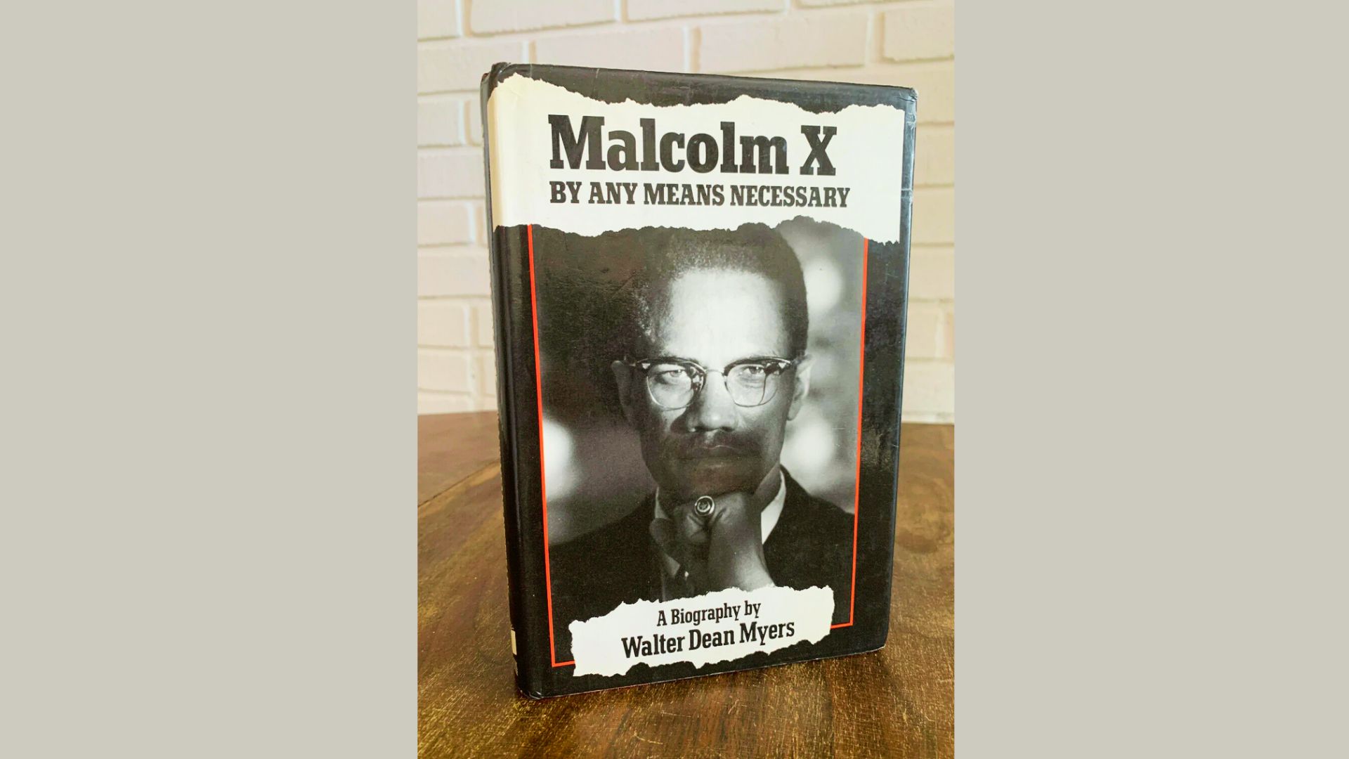 Sepia portrait of Malcolm X wearing glasses with his hand raised, overlaid with bold red and white title text.