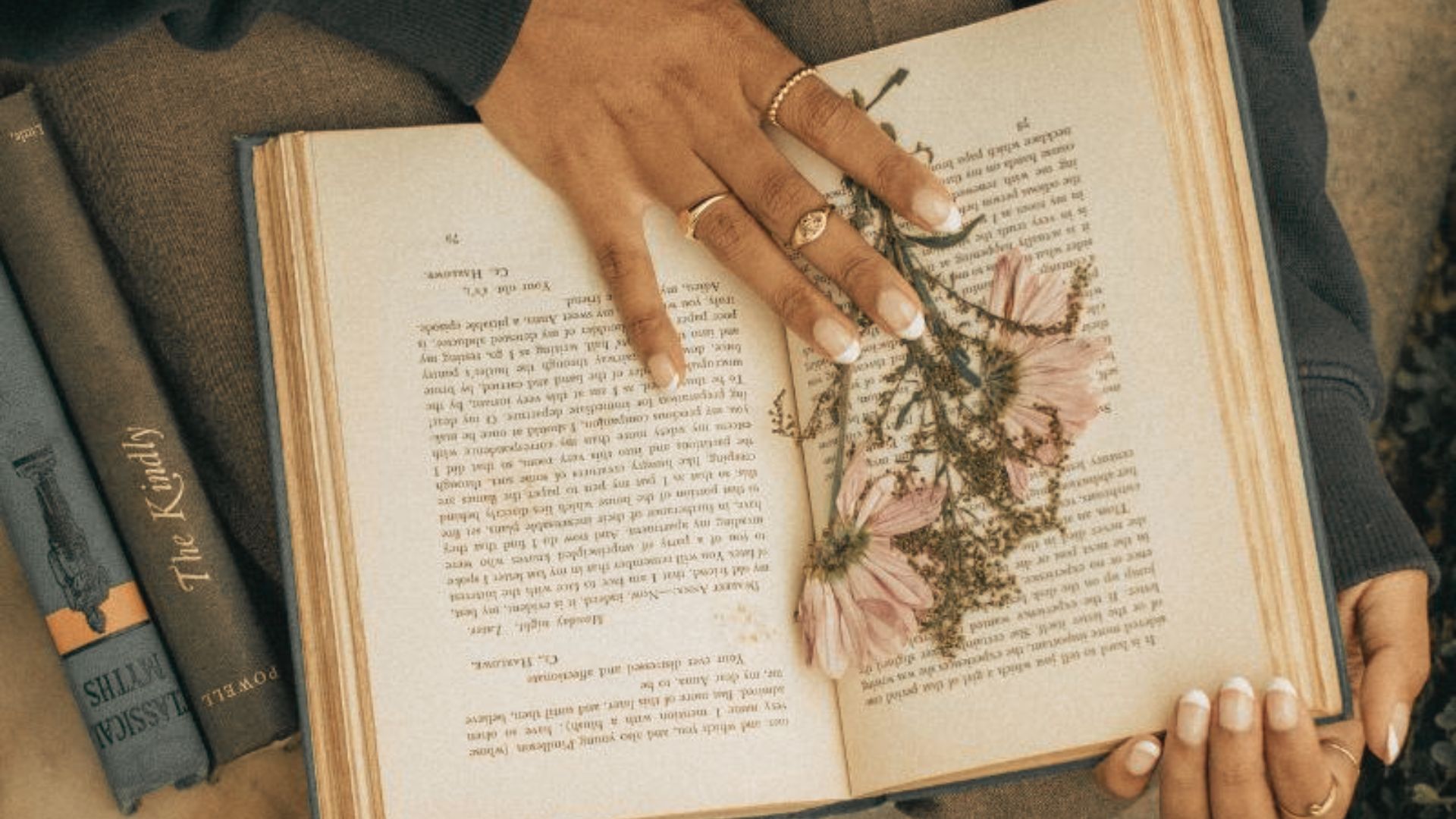 A hand adorned with rings rests on an open book, pressed pink flowers marking a favorite page.