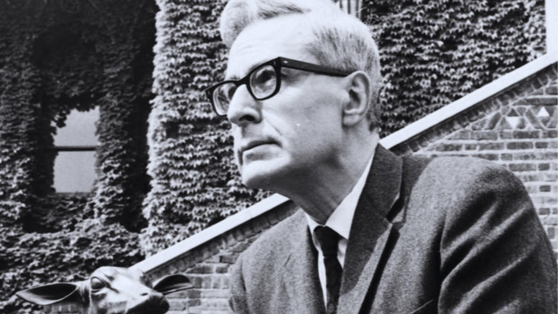 Black and white photo of man with glasses and suit beside ivy-covered brick building and sculpture.
