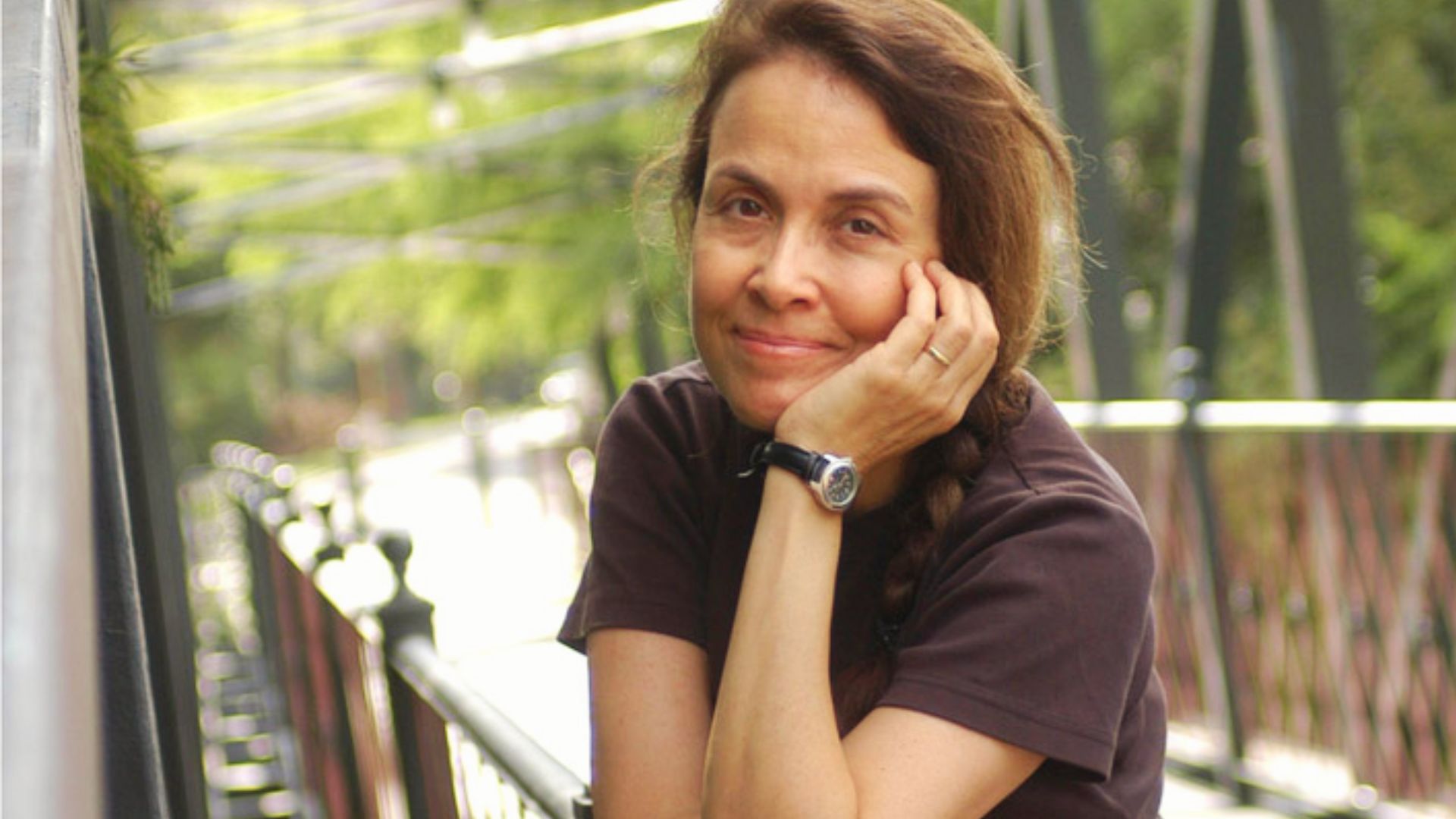 Poet Naomi Shihab Nye smiling outdoors on bridge, wearing brown shirt and wristwatch.