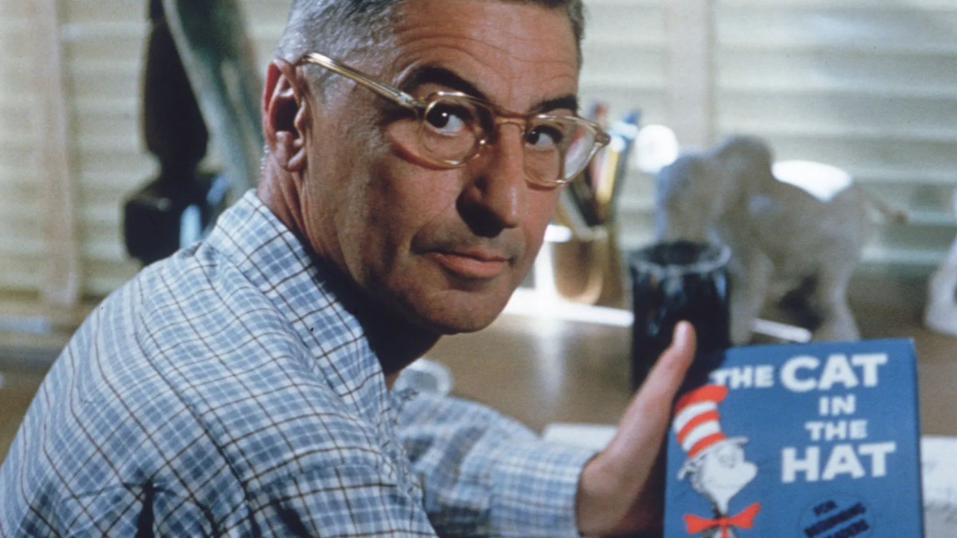Dr Seuss wearing glasses and a checkered shirt holding The Cat in the Hat book at his desk