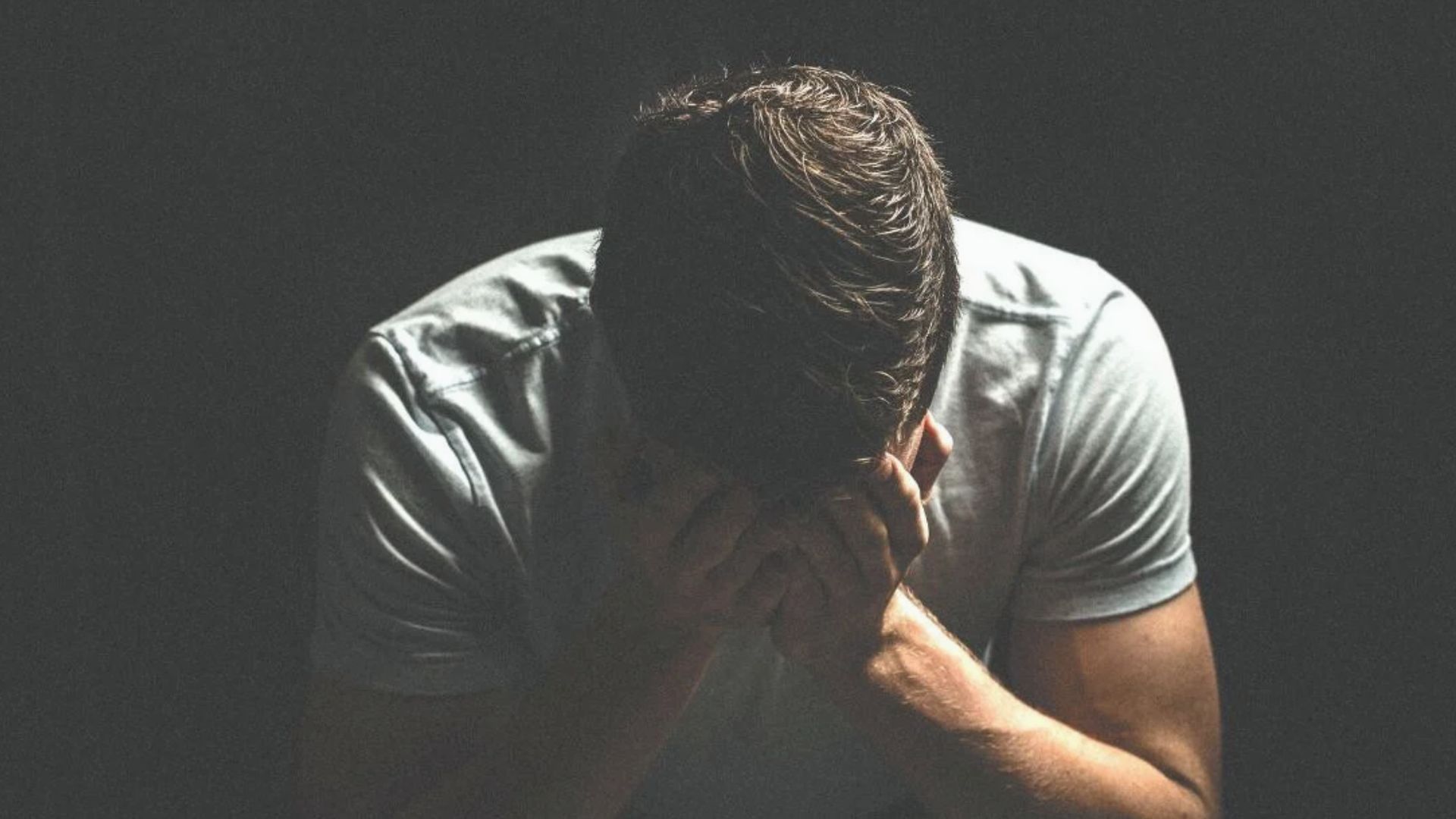 Man sitting alone with head bowed in hands against dark background