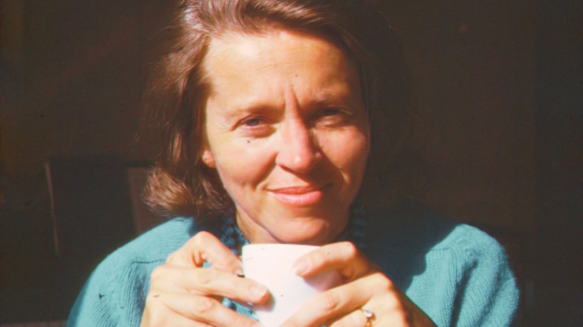 A woman smiling warmly while holding a white cup outdoors.