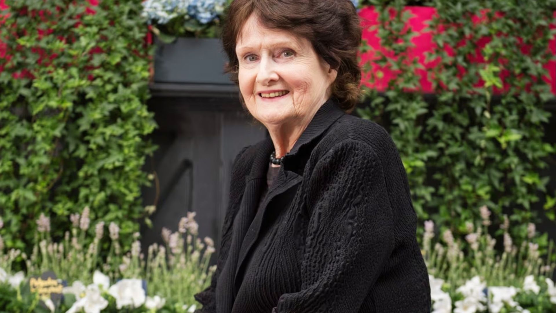 Eavan Boland smiling in a black jacket, standing before a lush garden with colourful flowers and foliage.