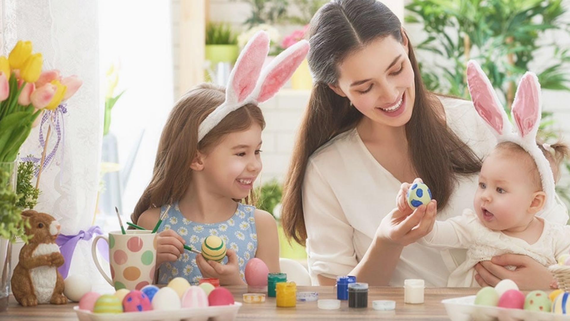 Mother with baby and daughter in bunny ears painting Easter eggs at home