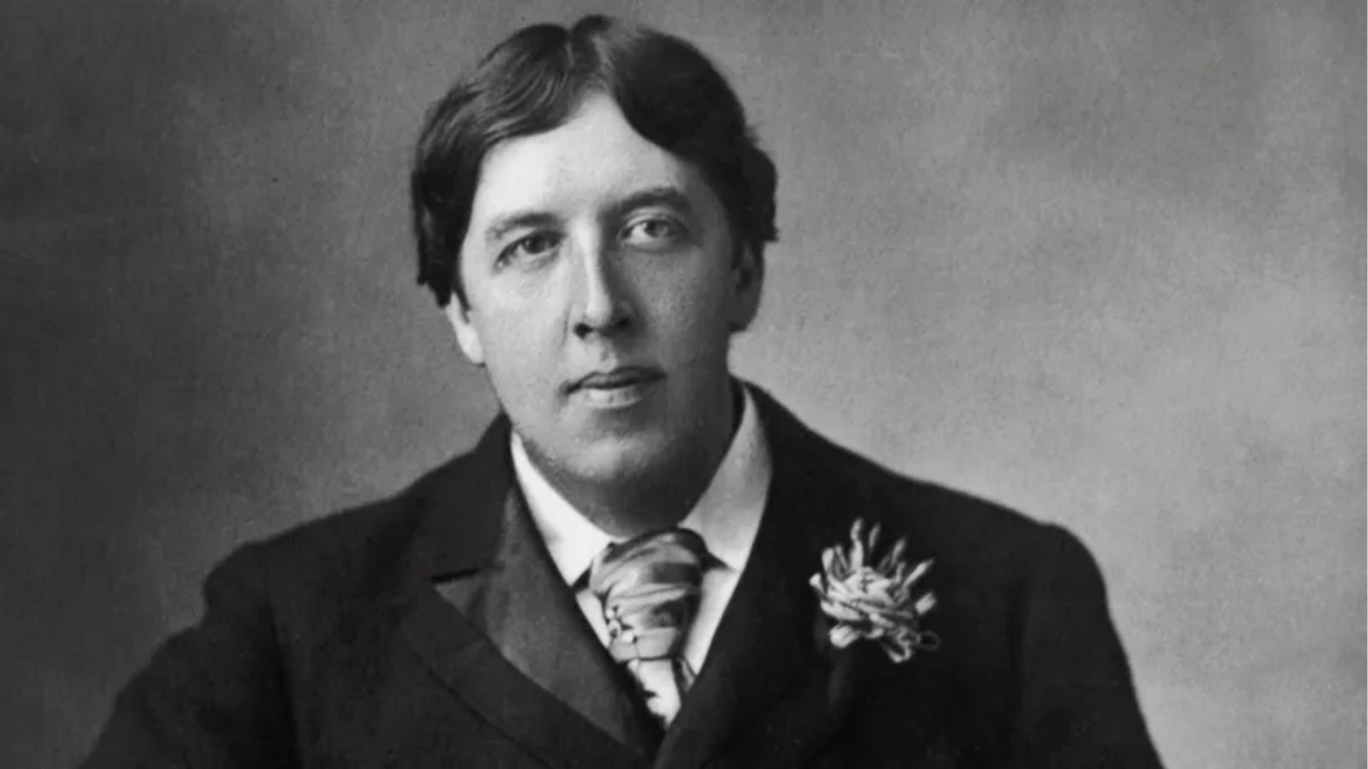Black-and-white portrait of Oscar Wilde in a dark suit, floral lapel pin, and cravat, gazing upward
