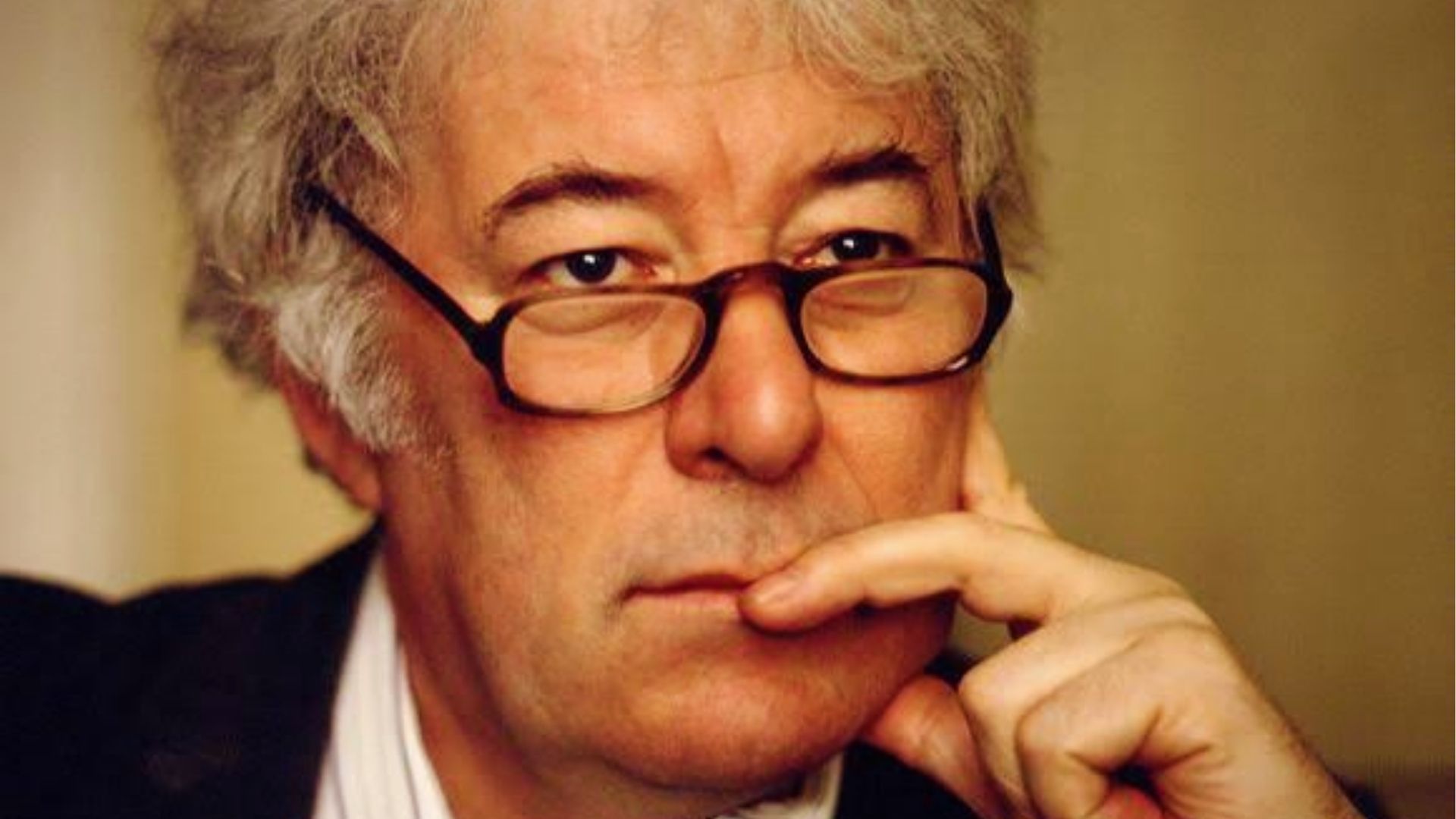 Close-up portrait of Seamus Heaney, white-haired man with dark glasses, resting chin on hand thoughtfully.