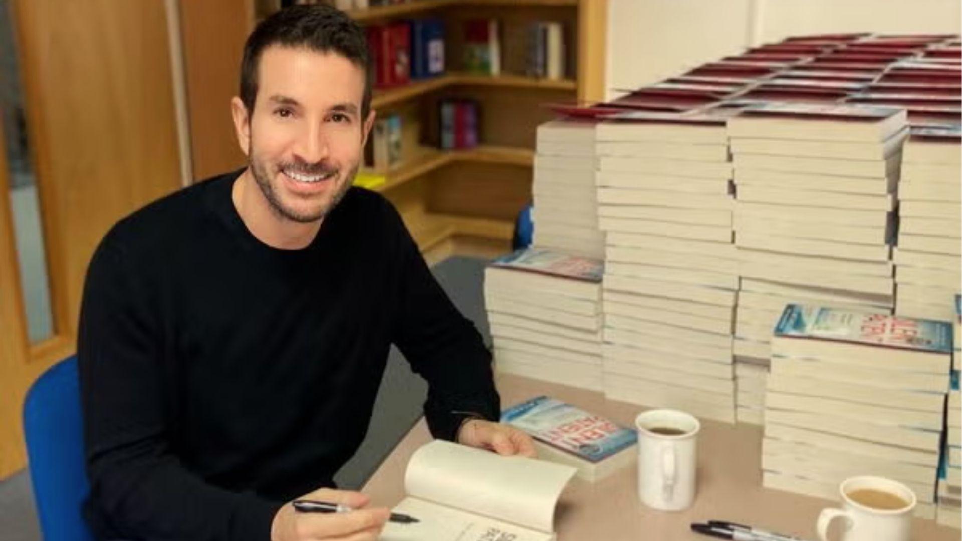 Alex Michaelides signing copies of books at a table surrounded by stacks of books