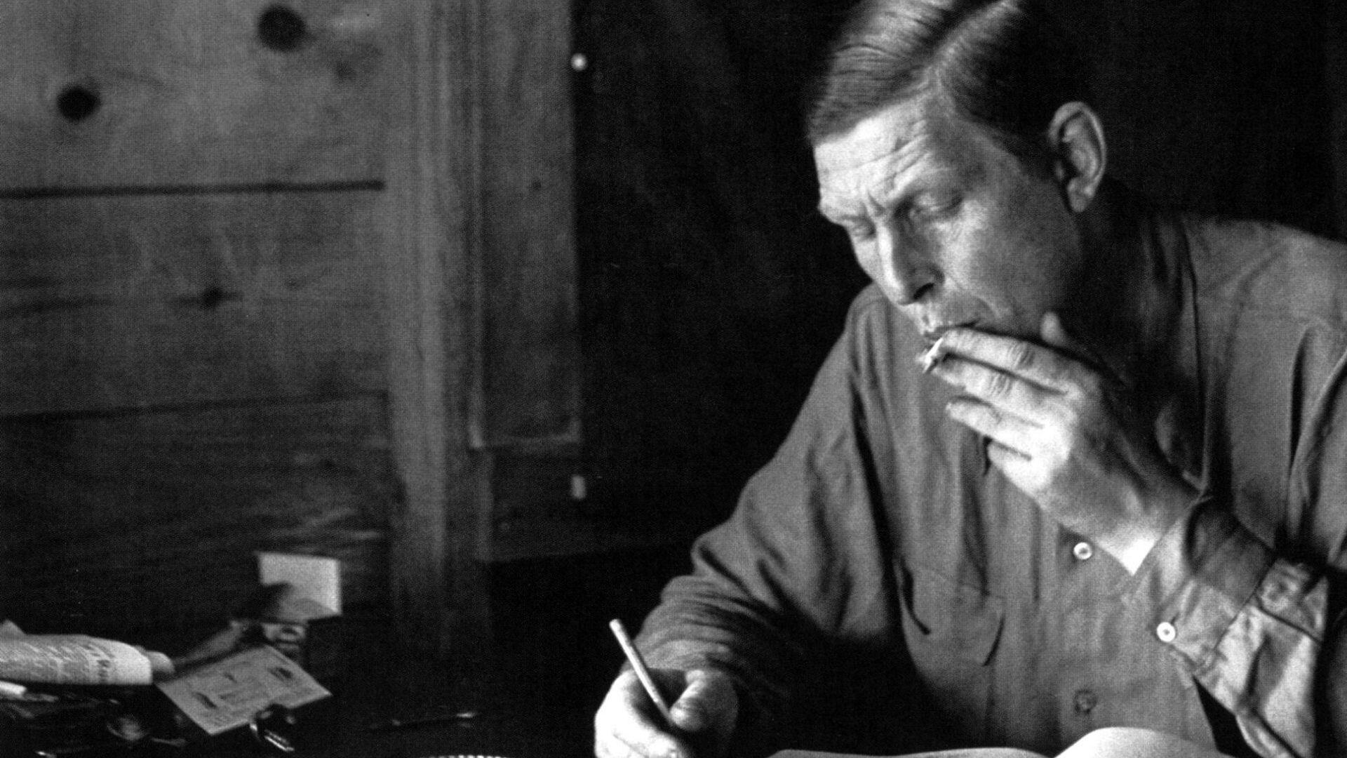 A man sits at a desk, holding a cigarette in one hand and writing with the other. The setting appears rustic, with wooden walls and minimal lighting. File name: black-and-white-photo-of-wh-auden-writing-at-desk