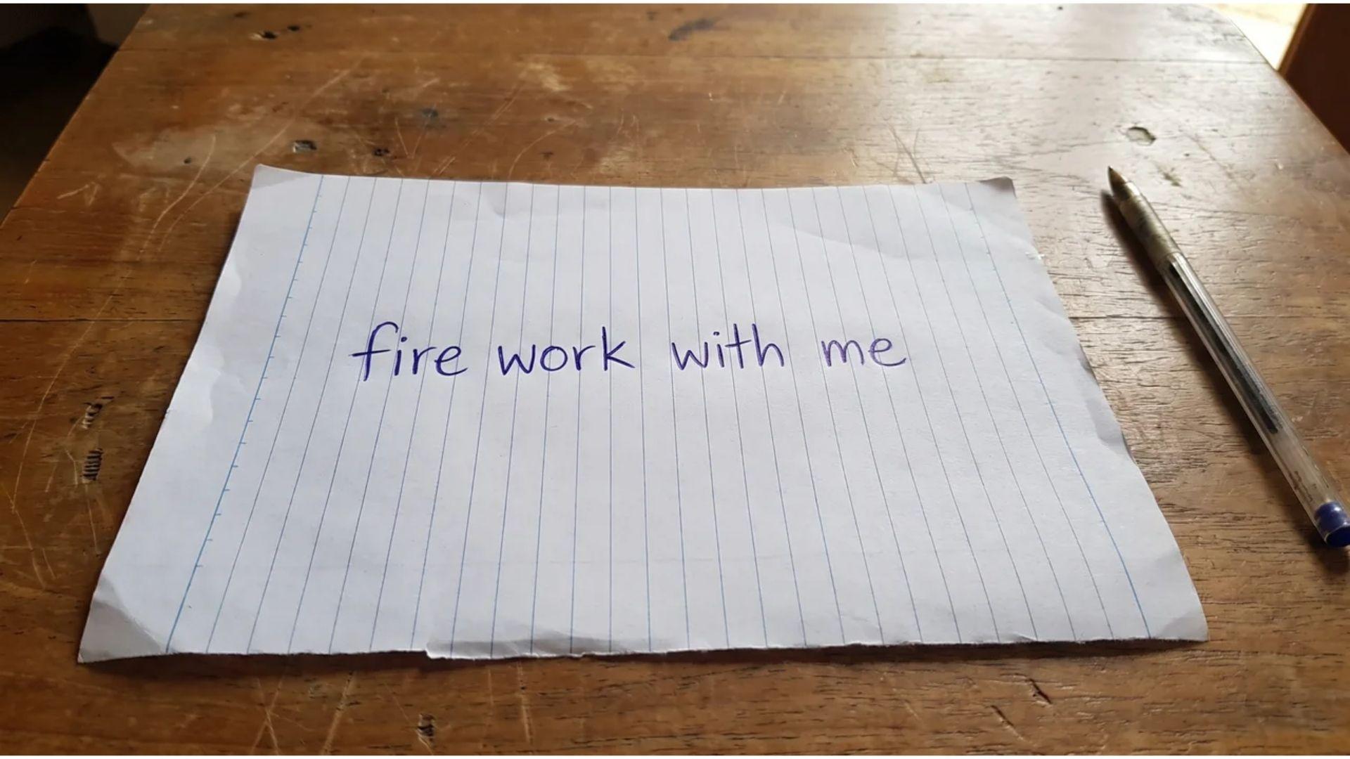 Handwritten note saying "fire work with me" on lined paper on a wooden table next to a blue pen.