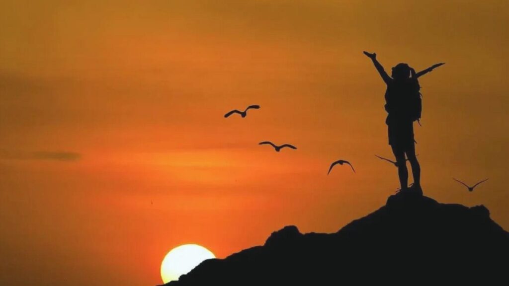 Person standing on rocky hilltop with arms raised against a vibrant sunset