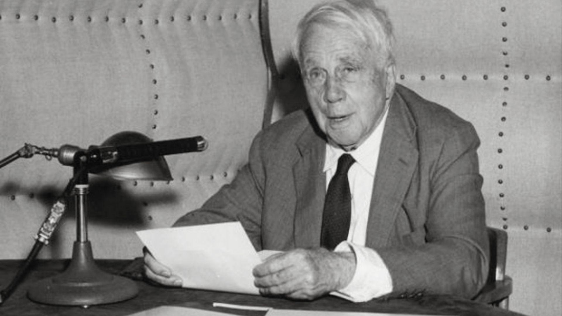 Elderly man in suit seated at desk holding papers near a microphone, black and white photo.