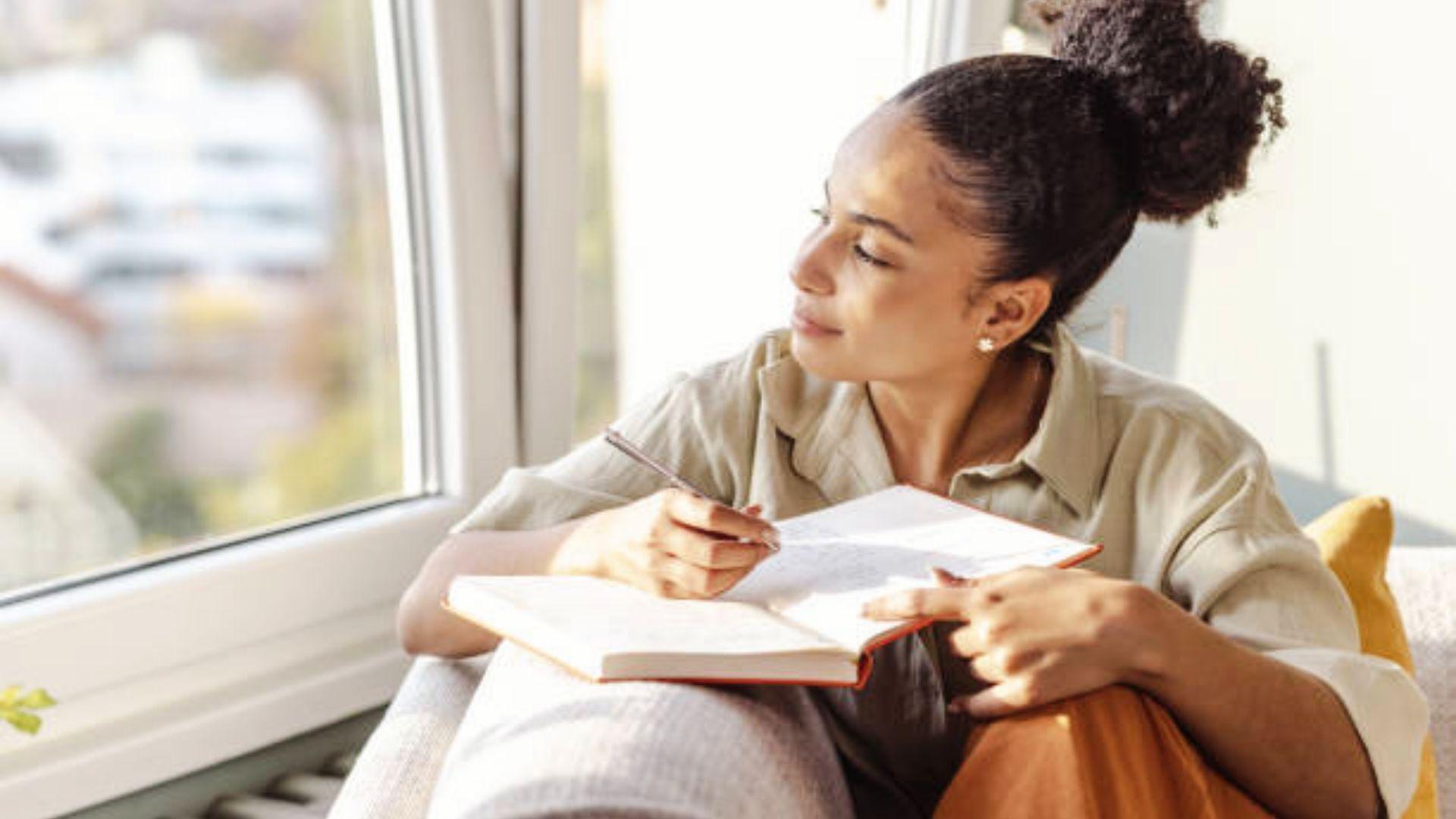 A woman writing in an open notebook with a pencil while sitting beside a window.File name-woman-writing-poem-in-notebook