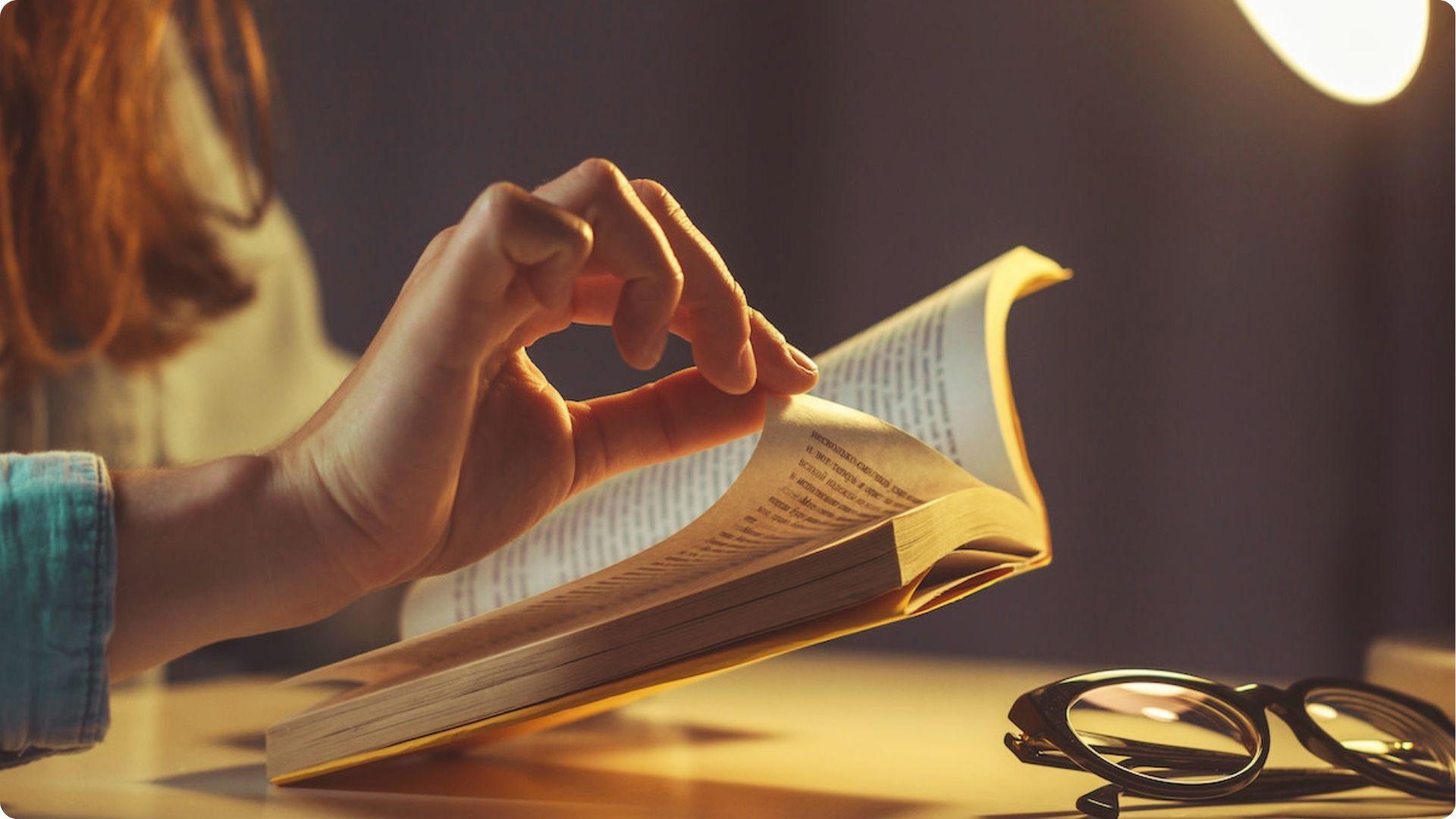 Woman's hand turning page of open book on desk with black glasses.File name: examples-of-motif-in-poetry