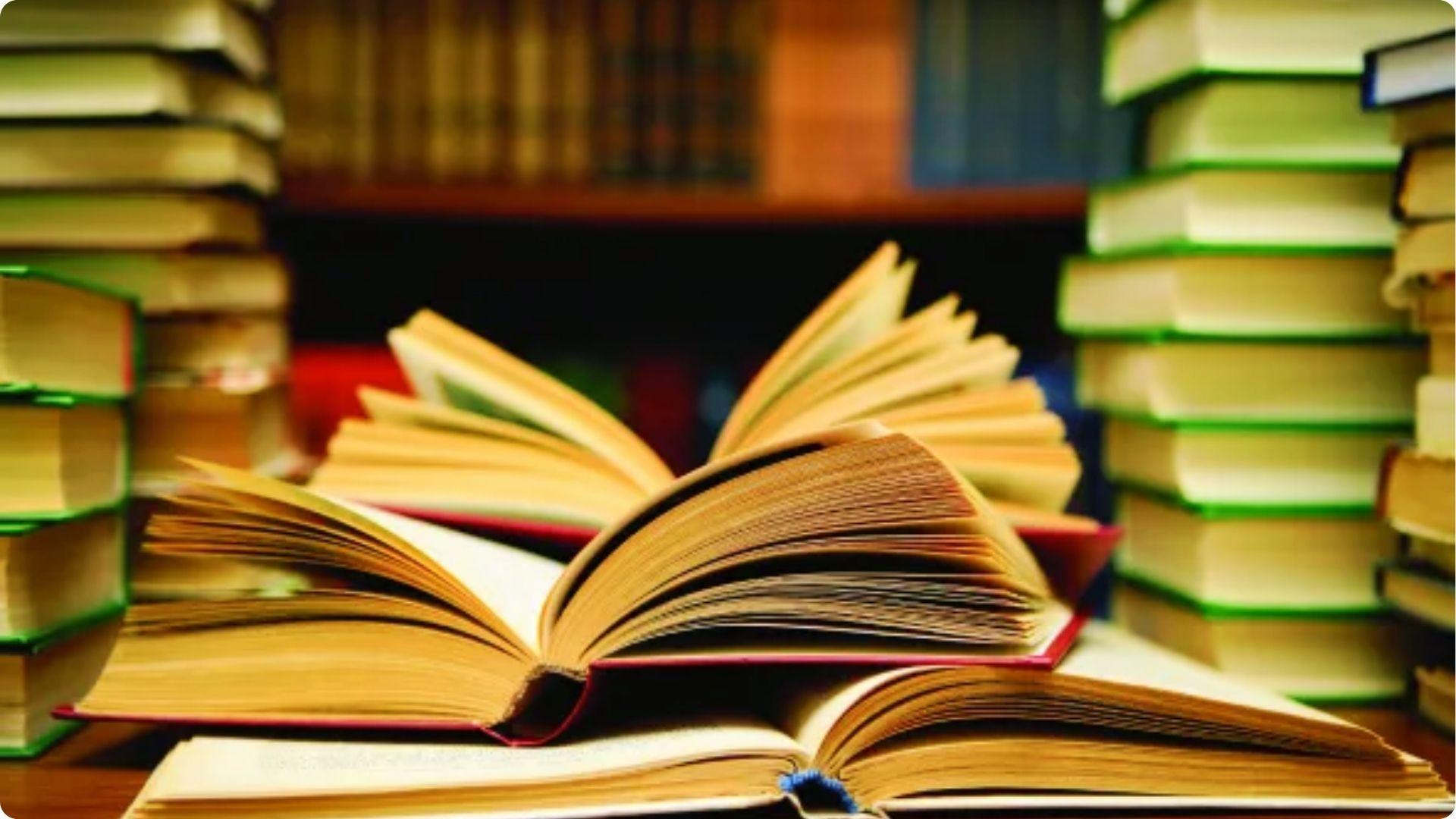Open books with fanned pages stacked on a table surrounded by tall piles of books.File name: example-of-motif-novels-play