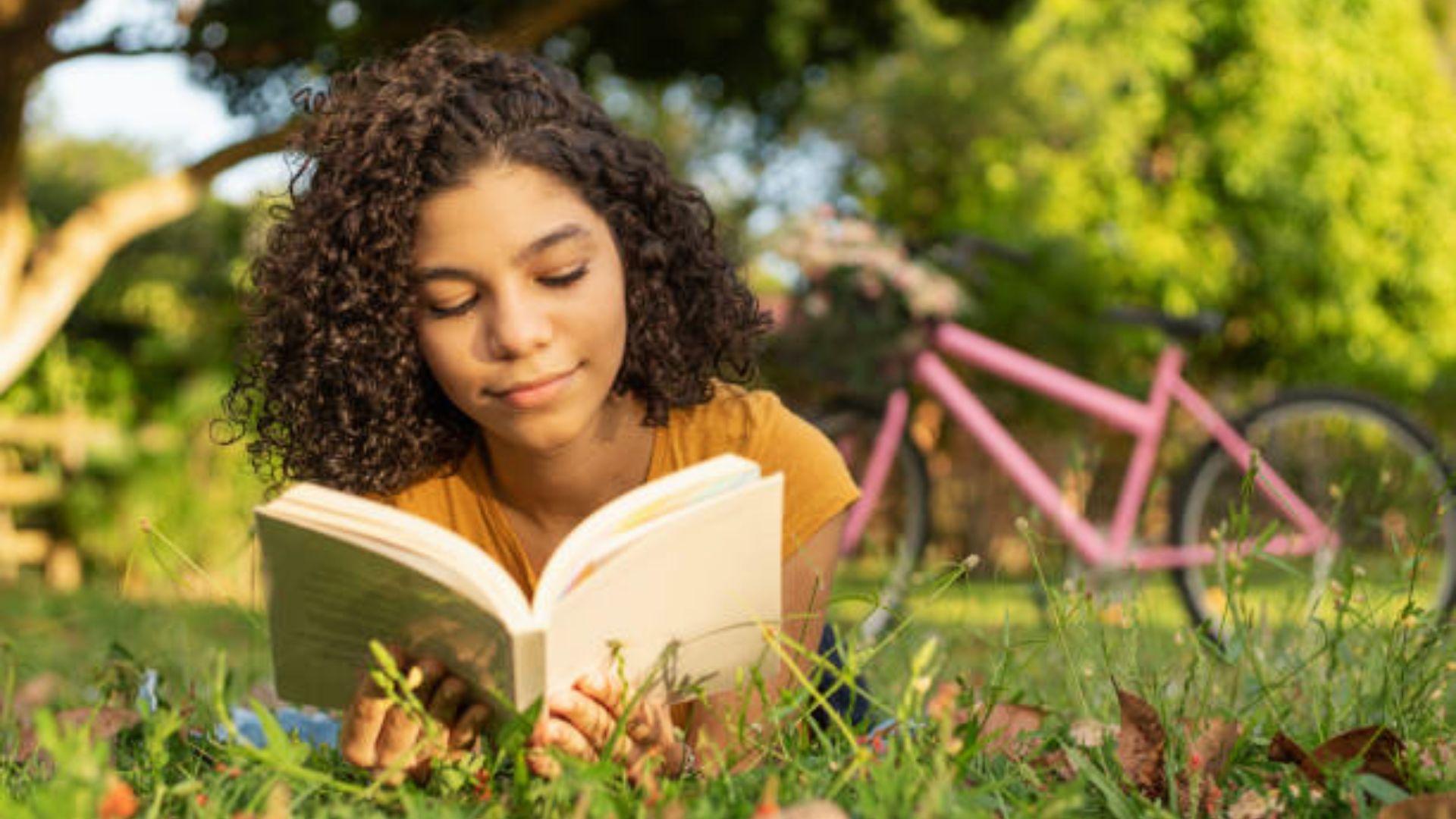 Girl reading an open book while lying on the grass in a sunny garden.File Name-girl-reading-book-outdoors