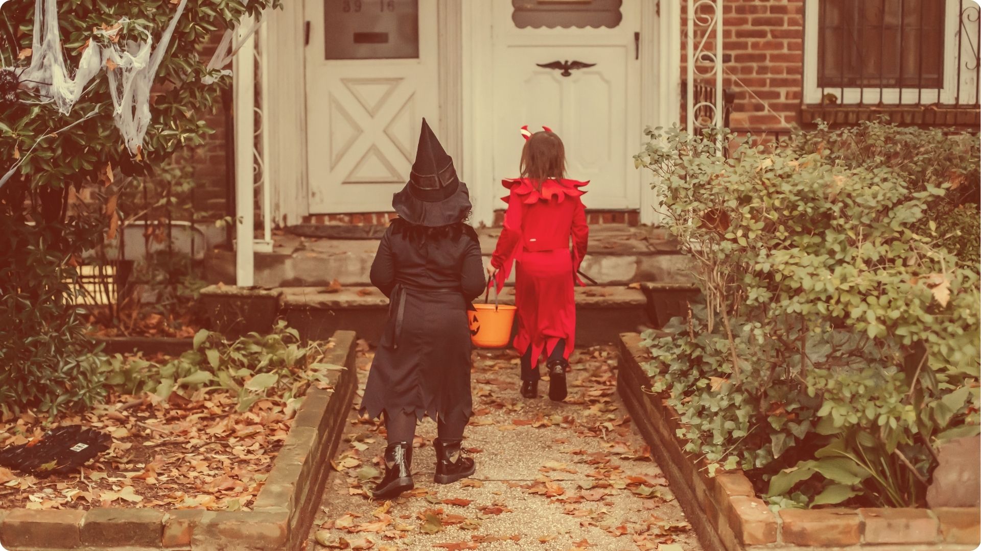Two small children in witch and devil costumes trick-or-treating with pumpkin buckets.