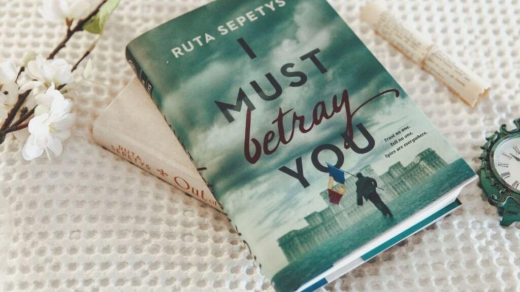 It Must Be Betrayal by Ruta Sepetys displayed with another book, flowers, and vintage clock.